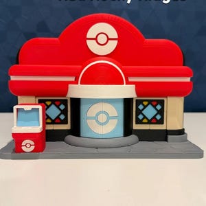 May include: A 3D-printed model of a building with the text "Red Rocky Ridges". The building is primarily red with white and blue accents. It features a central dome and a small vending machine-like structure on the side.