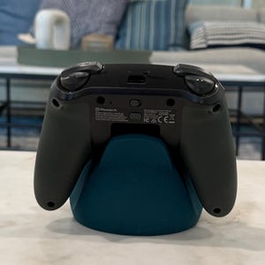 May include: A black and green video game controller on a teal stand. The controller features two black joysticks, a directional pad, and various buttons. The teal stand has a curved design. The controller rests on a white marble surface. The PowerA logo is visible.
