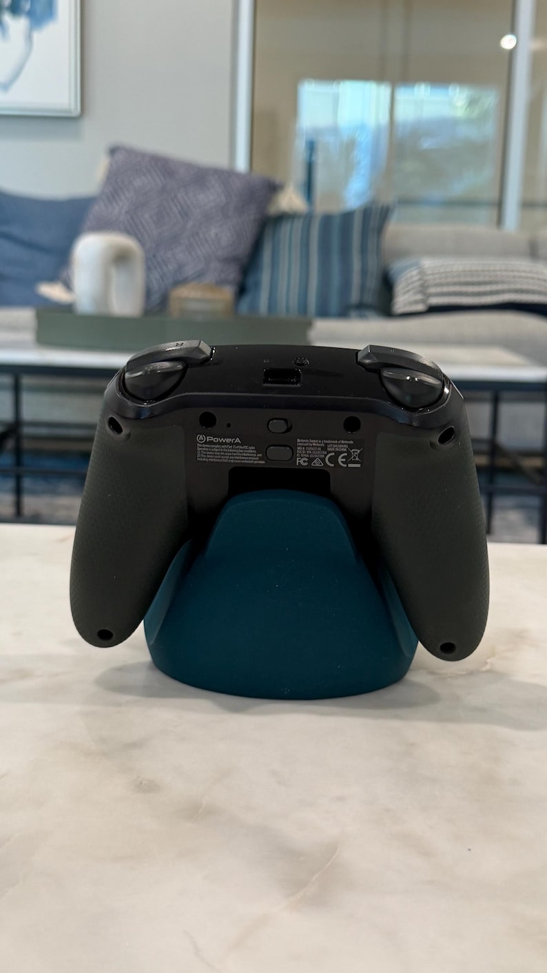 May include: A black and green video game controller on a teal stand. The controller features two black joysticks, a directional pad, and various buttons. The teal stand has a curved design. The controller rests on a white marble surface. The PowerA logo is visible.