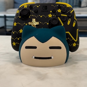 May include: A black and yellow Nintendo Switch controller with a star pattern and a gold cross button is displayed behind a Snorlax-shaped Switch dock. The dock is beige and teal, with a smiling face.