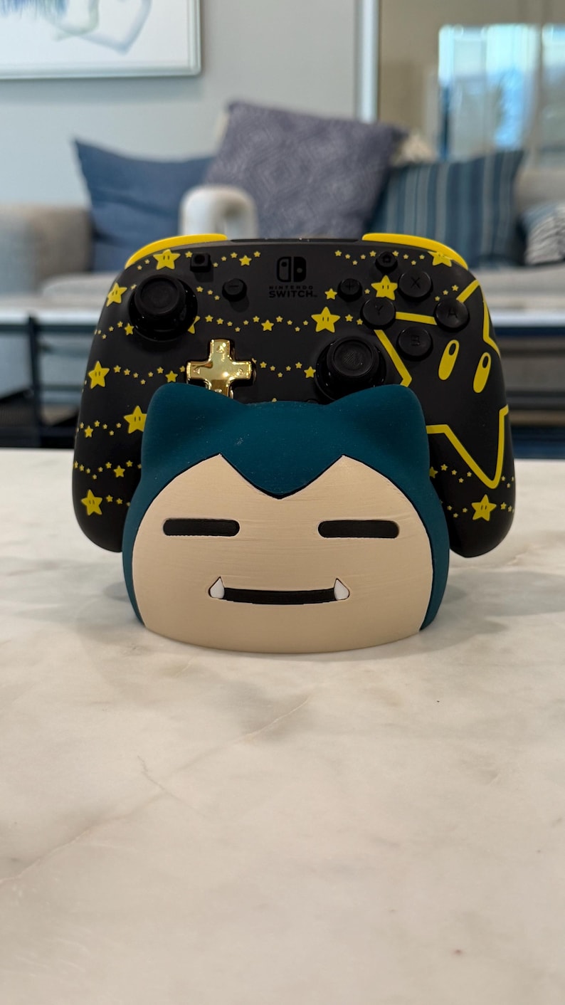 May include: A black and yellow Nintendo Switch controller with a star pattern and a gold cross button is displayed behind a Snorlax-shaped Switch dock. The dock is beige and teal, with a smiling face.