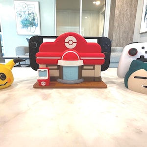 May include: A collection of Pok&eacute;mon-themed items, including a yellow Mimikyu plush, a red and white Pok&eacute;mon Center model, a Nintendo Switch, a white controller, and a Snorlax plush. The items are arranged on a marble surface.