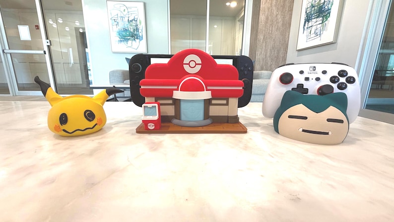 May include: A collection of Pok&eacute;mon-themed items, including a yellow Mimikyu plush, a red and white Pok&eacute;mon Center model, a Nintendo Switch, a white controller, and a Snorlax plush. The items are arranged on a marble surface.