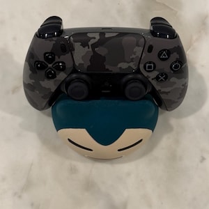 May include: A black and grey camouflage-patterned video game controller sits on a teal and cream-coloured character-shaped accessory. The controller has black buttons and directional controls.