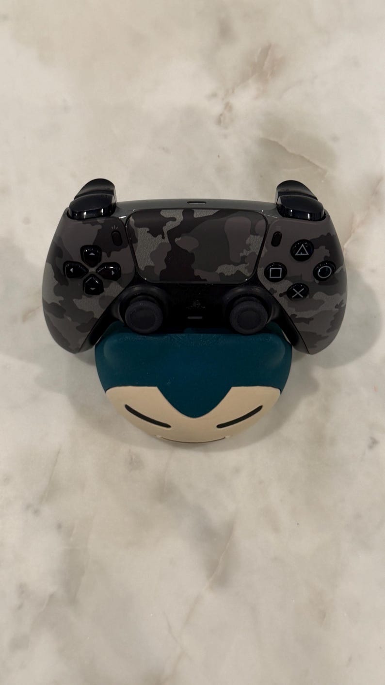 May include: A black and grey camouflage-patterned video game controller sits on a teal and cream-coloured character-shaped accessory. The controller has black buttons and directional controls.