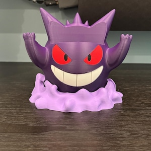 Pode incluir: Uma figura Gengar roxa impressa em 3D, com olhos vermelhos e um sorriso largo. A figura est&aacute; posada com os bra&ccedil;os levantados e est&aacute; sobre uma base de nuvem lavanda. Um item colecion&aacute;vel divertido.