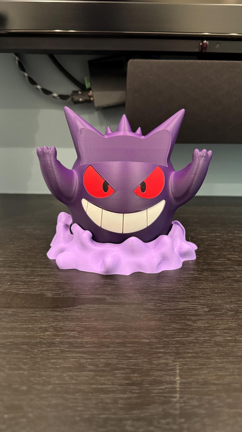 Pode incluir: Uma figura Gengar roxa impressa em 3D, com olhos vermelhos e um sorriso largo. A figura est&aacute; posada com os bra&ccedil;os levantados e est&aacute; sobre uma base de nuvem lavanda. Um item colecion&aacute;vel divertido.