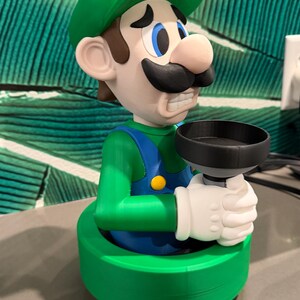 May include: A 3D-printed figurine of Luigi from the Super Mario Bros. series. The figure is wearing a green hat and overalls, with a black mustache and white gloves, holding a black plunger. The base is green.