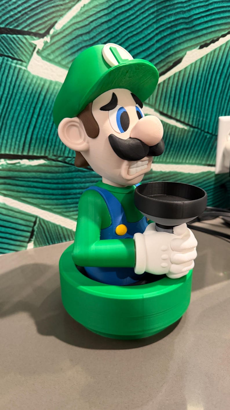 May include: A 3D-printed figurine of Luigi from the Super Mario Bros. series. The figure is wearing a green hat and overalls, with a black mustache and white gloves, holding a black plunger. The base is green.