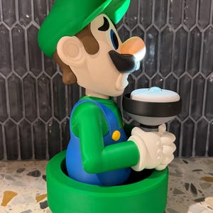 May include: A 3D-printed figurine of Luigi from the Super Mario Bros. series. The figure is wearing a green hat, green shirt, blue overalls, and white gloves. He is holding a white and black object. The figurine is set on a green base.