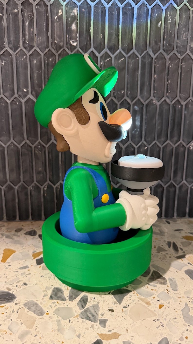 May include: A 3D-printed figurine of Luigi from the Super Mario Bros. series. The figure is wearing a green hat, green shirt, blue overalls, and white gloves. He is holding a white and black object. The figurine is set on a green base.