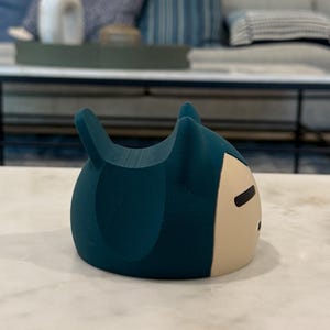 May include: A teal-coloured, cartoon-style figurine with a rounded body and two pointed ears. The front of the figurine is a light beige colour with a simple black facial expression. The figurine is sitting on a white marble surface.