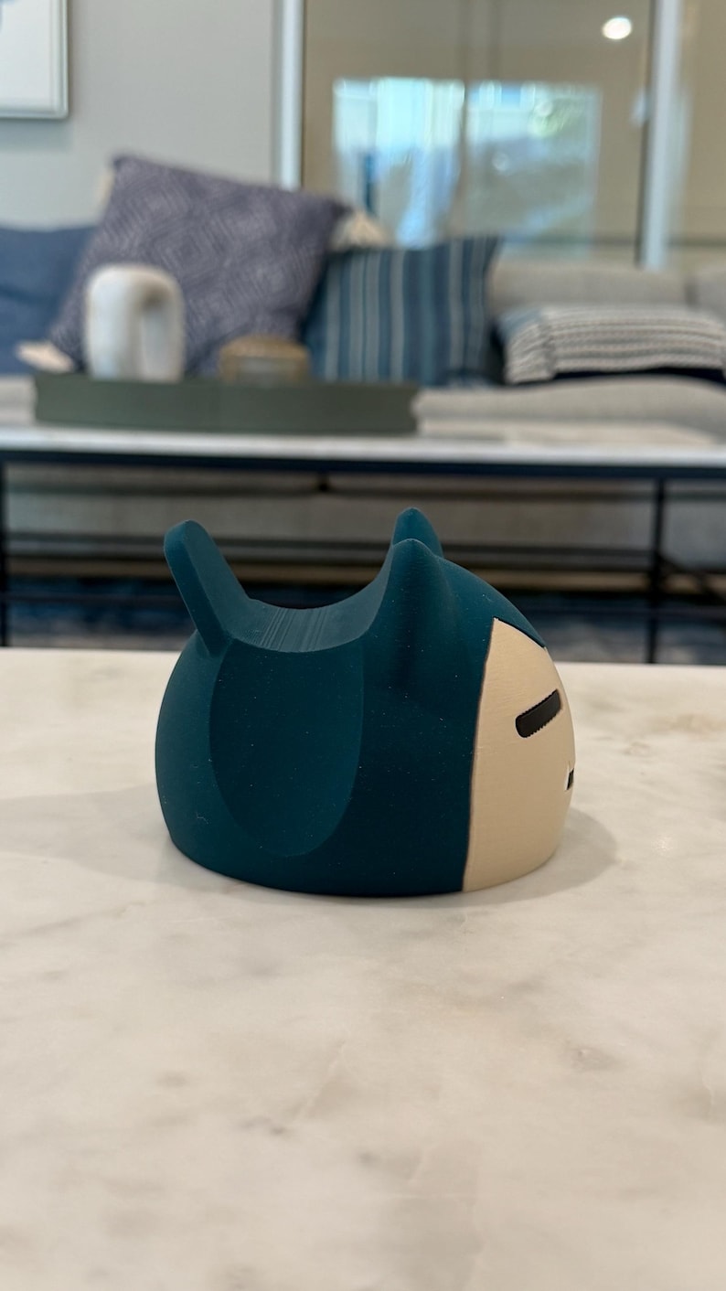 May include: A teal-coloured, cartoon-style figurine with a rounded body and two pointed ears. The front of the figurine is a light beige colour with a simple black facial expression. The figurine is sitting on a white marble surface.