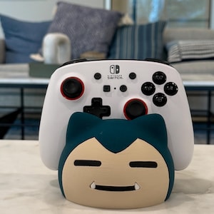 May include: A white Nintendo Switch controller with red detailing and black buttons sits above a blue and tan Snorlax-shaped stand. The controller has a black directional pad and the Nintendo Switch logo.