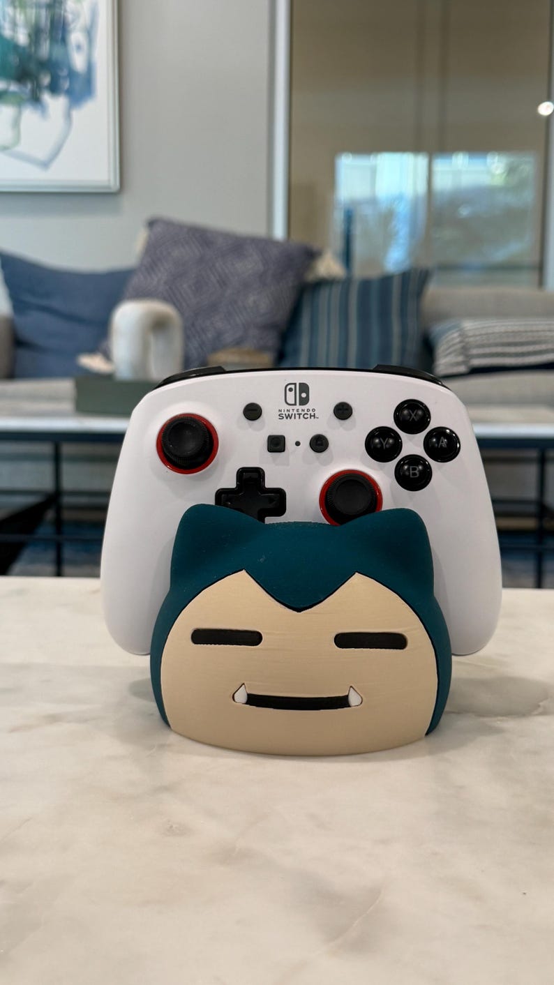 May include: A white Nintendo Switch controller with red detailing and black buttons sits above a blue and tan Snorlax-shaped stand. The controller has a black directional pad and the Nintendo Switch logo.