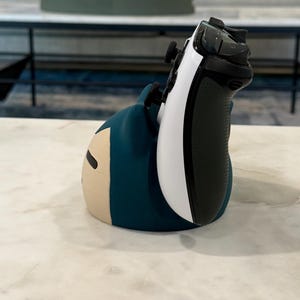 May include: A blue and beige character-shaped controller holder with a white and black game controller. The holder is on a white marble surface. The controller is white and black.