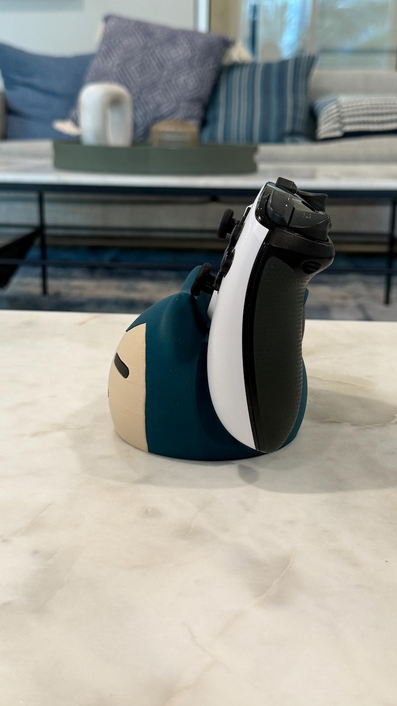 May include: A blue and beige character-shaped controller holder with a white and black game controller. The holder is on a white marble surface. The controller is white and black.