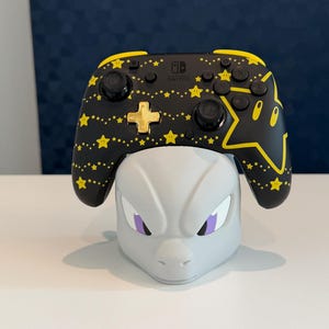 Puede incluir: Un mando Nintendo Switch Pro negro con un patrón de estrellas amarillas y una cruceta dorada, apoyado sobre un soporte gris claro con forma de cabeza de personaje y ojos morados. El mando incluye botones estándar, joysticks y el logo de la marca.
