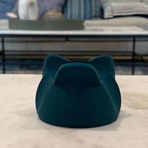 May include: A teal-coloured, cat-shaped object. The object has a rounded base and four curved sections that form the cat's ears. It sits on a white and grey marble surface. The background is blurred, showing a living room setting.