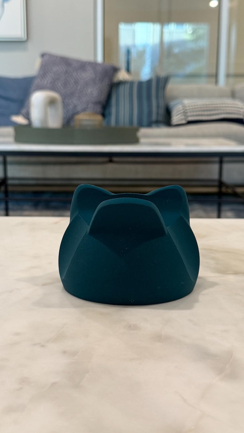 May include: A teal-coloured, cat-shaped object. The object has a rounded base and four curved sections that form the cat's ears. It sits on a white and grey marble surface. The background is blurred, showing a living room setting.
