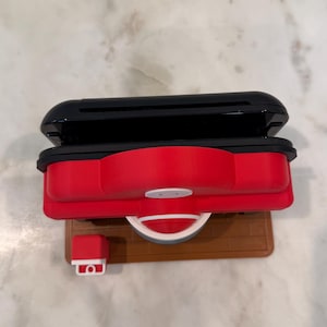 May include: A red and black toy grill with a white handle and a small red and white accessory. The grill sits on a brown base. The top of the grill is black.