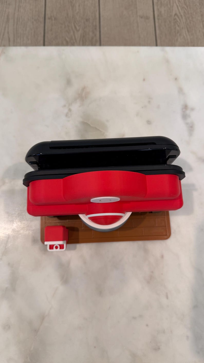 May include: A red and black toy grill with a white handle and a small red and white accessory. The grill sits on a brown base. The top of the grill is black.