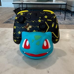 May include: A black Nintendo Switch controller with yellow star accents and a gold cross-shaped button is displayed behind a blue Bulbasaur-shaped accessory. The Bulbasaur has red eyes and green spots. The items are on a white marble surface.