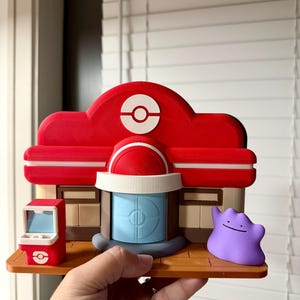 May include: A 3D-printed model of a Pok&eacute;mon Center, featuring a red roof with a white Pok&eacute; Ball symbol, a blue door, and a small red arcade machine. A purple Ditto figure stands nearby. The model is held in a hand.
