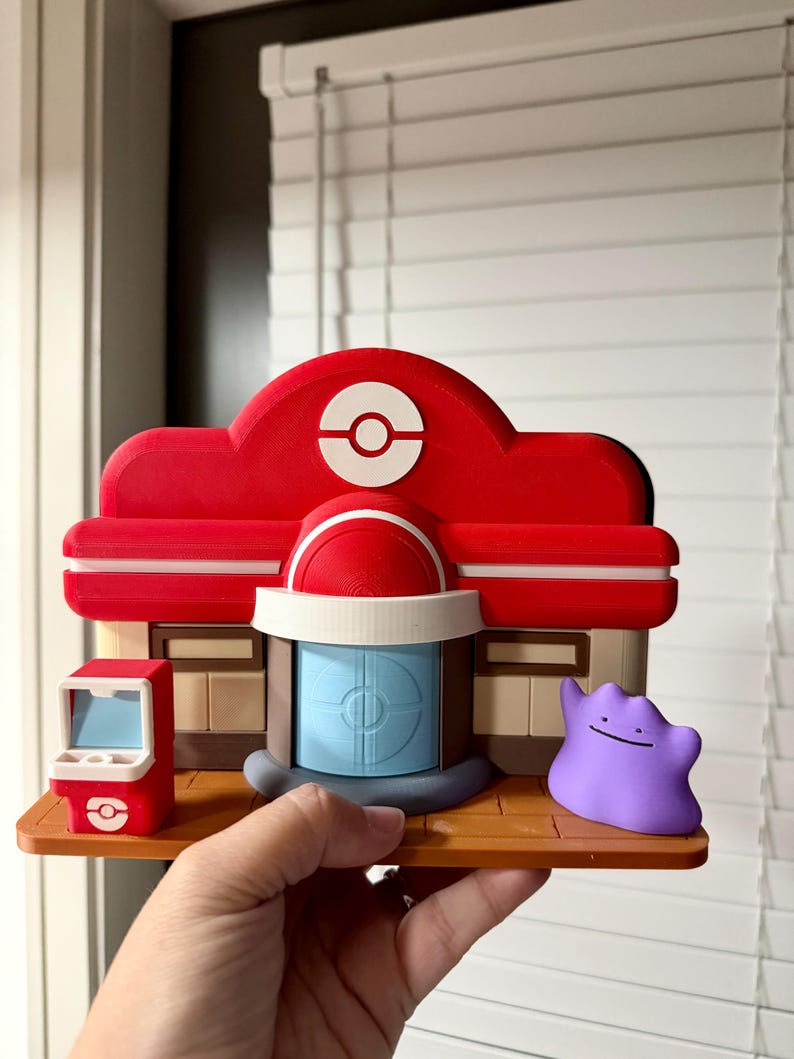 May include: A 3D-printed model of a Pok&eacute;mon Center, featuring a red roof with a white Pok&eacute; Ball symbol, a blue door, and a small red arcade machine. A purple Ditto figure stands nearby. The model is held in a hand.