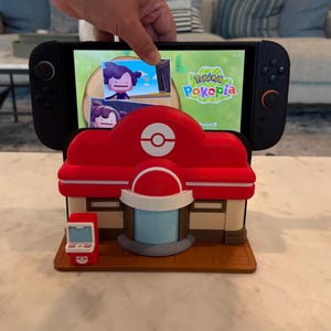 May include: A red and white Pokemon Center-themed Nintendo Switch stand. The stand features a detailed design of the iconic building, with a matching red and white PokeStop on the side. The Switch displays the game "Pok&eacute;pia".