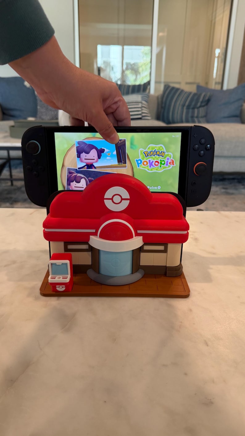 May include: A red and white Pokemon Center-themed Nintendo Switch stand. The stand features a detailed design of the iconic building, with a matching red and white PokeStop on the side. The Switch displays the game "Pok&eacute;pia".