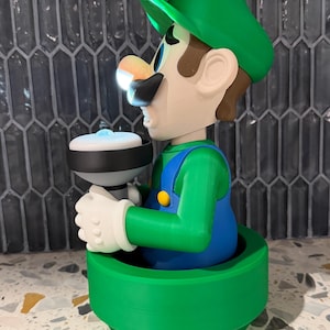 May include: A 3D-printed figurine of Luigi from the Super Mario Bros. series. The figure is wearing a green hat, blue overalls, and white gloves, holding a white and black object. The base is green, and the figure has a light shining from the object.
