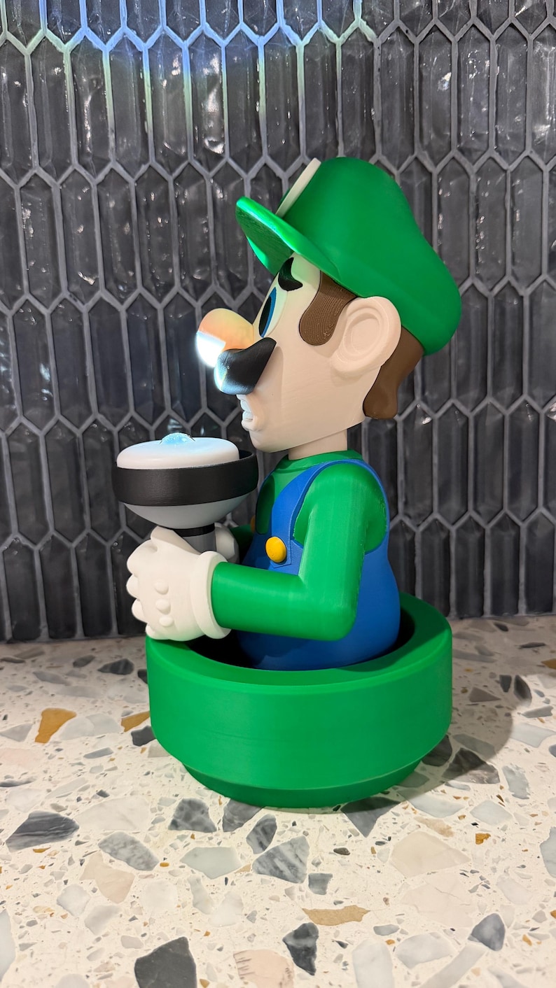May include: A 3D-printed figurine of Luigi from the Super Mario Bros. series. The figure is wearing a green hat, blue overalls, and white gloves, holding a white and black object. The base is green, and the figure has a light shining from the object.