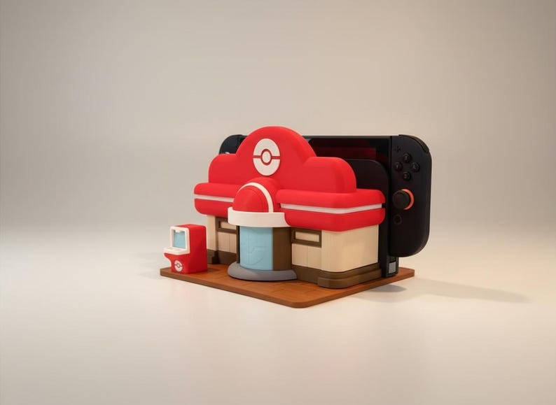 May include: A red and white themed Nintendo Switch dock shaped like a Pok&eacute;mon Center. The dock has a small red arcade machine and a brown base. A black Nintendo Switch is inserted into the dock.