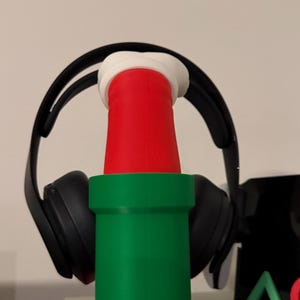 以下が含まれることがあります： 緑と赤のパイプの形をした3Dプリントのヘッドフォンスタンド。白い上部が付いています。黒いヘッドフォンがスタンドに置かれています。スタンドは赤いベースにあります。デザインはビデオゲームを連想させます。