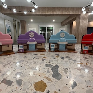May include: Four colorful, miniature structures resembling a shop or building, each with a different color scheme: pink, purple, blue, and red. Each structure has a rounded roof, a central circular design, and a light blue counter.