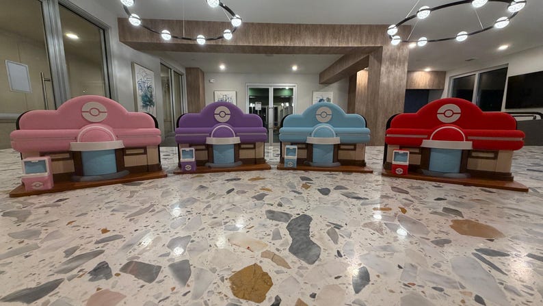 May include: Four colorful, miniature structures resembling a shop or building, each with a different color scheme: pink, purple, blue, and red. Each structure has a rounded roof, a central circular design, and a light blue counter.