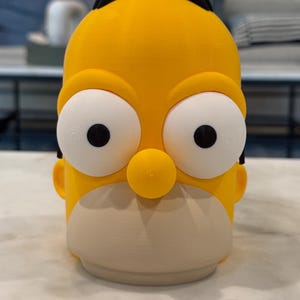 May include: A yellow, 3D-printed figurine of a cartoon character's head, featuring large white eyes with black pupils, a tan face, and a black band on top. The figurine is on a white surface.