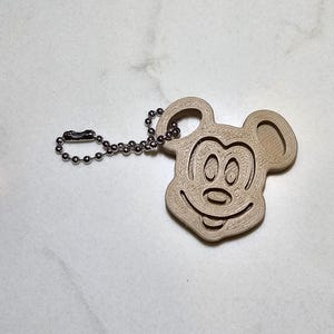 Könnte beinhalten: Ein hellbrauner Mickey Mouse-Schlüsselanhänger mit einer silbernen Kugelkette. Der Schlüsselanhänger besteht aus einem hellfarbigen Material und zeigt eine detaillierte Umrisszeichnung von Mickey Mouses Gesicht, einschließlich Ohren, Augen, Nase und Mund.