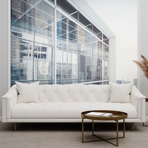 Puede incluir: Un sofá de cuero blanco con tapicería capitoné y dos almohadas se encuentra frente a una ventana grande con vista a un horizonte urbano. Una mesa de café redonda de metal dorado con una revista encima se encuentra frente al sofá. Un jarrón alto con hierba de la pampa seca se encuentra sobre una mesa auxiliar de metal dorado más pequeña.