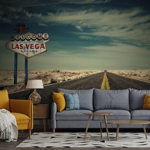 Könnte beinhalten: Ein Wohnzimmer-Interieur mit einem grauen Sofa, zwei gelben Sesseln und zwei Couchtischen. Die Wand hinter den Möbeln zeigt ein Wandbild einer Wüstenstraße, die zu einem Vintage-Schild "Welcome to Fabulous Las Vegas, Nevada" führt.