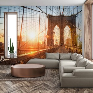 May include: A living room interior design with a large wall mural featuring a sunset view of the Brooklyn Bridge in New York City. A light gray sectional sofa with multiple pillows is positioned in front of the mural. A round, wooden coffee table sits in front of the sofa.