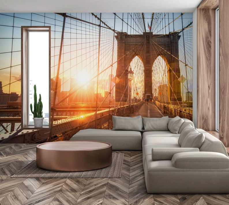 May include: A living room interior design with a large wall mural featuring a sunset view of the Brooklyn Bridge in New York City. A light gray sectional sofa with multiple pillows is positioned in front of the mural. A round, wooden coffee table sits in front of the sofa.
