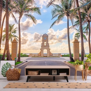 Puede incluir: Una escena exterior escénica con una torre del reloj, palmeras y una vista costera. Un banco bajo de madera con patas negras, una cesta tejida y una alfombra con lunares negros están en primer plano. También se ven una impresión botánica enmarcada y un bolso tote tejido.