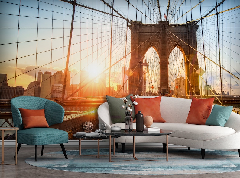 May include: A living room interior design featuring a white couch with orange and teal throw pillows, a teal armchair with an orange pillow, two black coffee tables, and a wall mural of the Brooklyn Bridge at sunset.