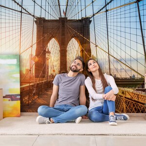 May include: A couple sits on a rug in front of a wall mural depicting the Brooklyn Bridge. The couple is looking up at the bridge. There are boxes and other items in the room, suggesting they are moving into a new home.