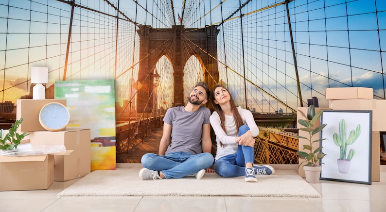May include: A couple sits on a rug in front of a wall mural depicting the Brooklyn Bridge. The couple is looking up at the bridge. There are boxes and other items in the room, suggesting they are moving into a new home.