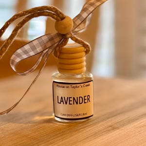 May include: A small glass car air freshener bottle with a wooden cap and a gingham ribbon. The label reads "LAVENDER" and "CAR DIFFUSER | 8ml". The bottle is suspended by a braided cord.