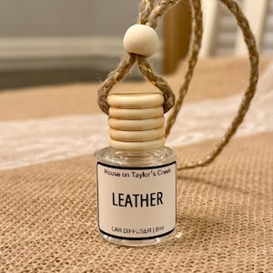 May include: A small, clear glass car diffuser bottle with a wooden cap and a rope for hanging. The label reads "LEATHER" and "CAR DIFFUSER | 8ml". The bottle is suspended against a neutral background.