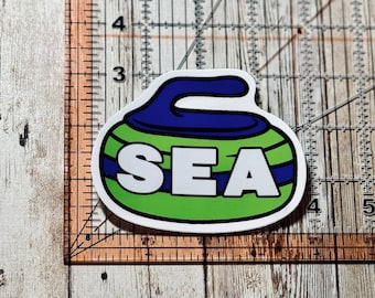 May include: A sticker featuring a curling stone design with the word "SEA" in white block letters. The stone has a green body with blue and white stripes and a blue handle. The sticker is on a wooden surface.